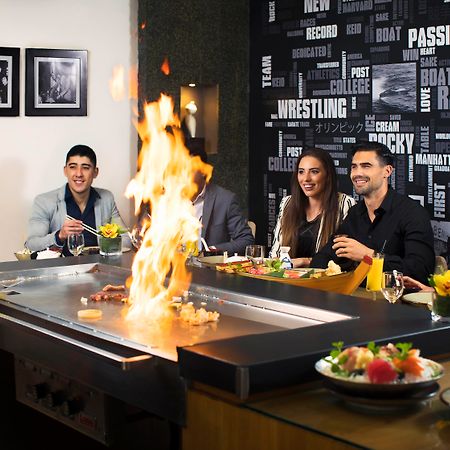 Amwaj Rotana, Jumeirah Beach - Dubai Esterno foto The photo shows a lively dining scene at a restaurant where hibachi cooking is taking place. In the foreground, a chef is preparing food on a grill, creating flames, which adds excitement to the experience. There are five diners seated around the gri