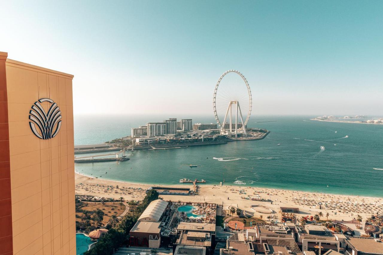 Amwaj Rotana, Jumeirah Beach - Dubai Esterno foto The photo shows a stunning coastal view featuring a large Ferris wheel, likely situated on an island or seaside area. In the foreground, there is a high-rise building with a distinctive logo, while below, you can see a beach with umbrellas and people