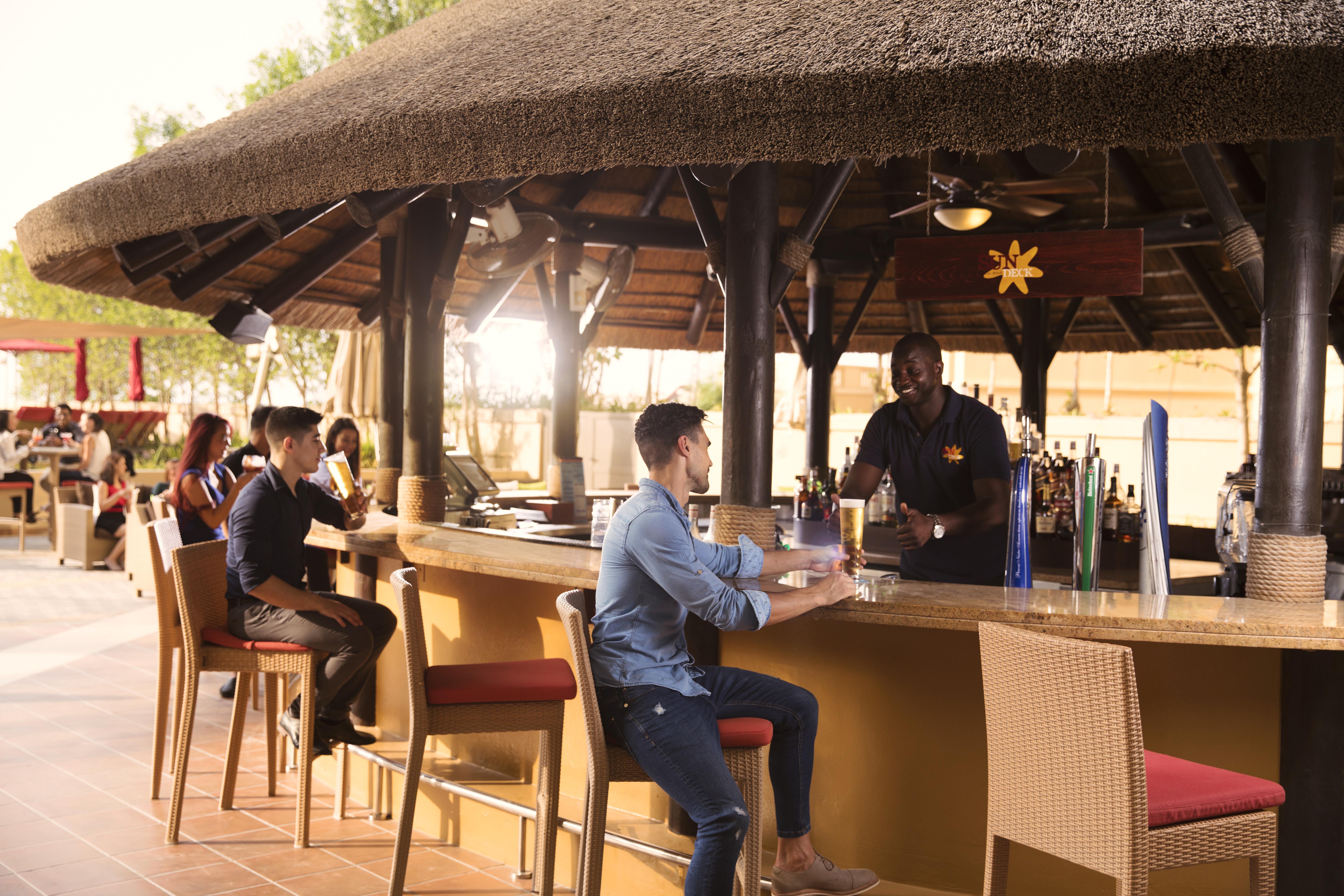 Amwaj Rotana, Jumeirah Beach - Dubai Esterno foto The photo shows a bar area with a thatched roof. There are several people seated at the bar. On the left, two individuals are chatting together, while a bartender is serving a customer in the foreground. The customer is wearing a blue shirt and sitti