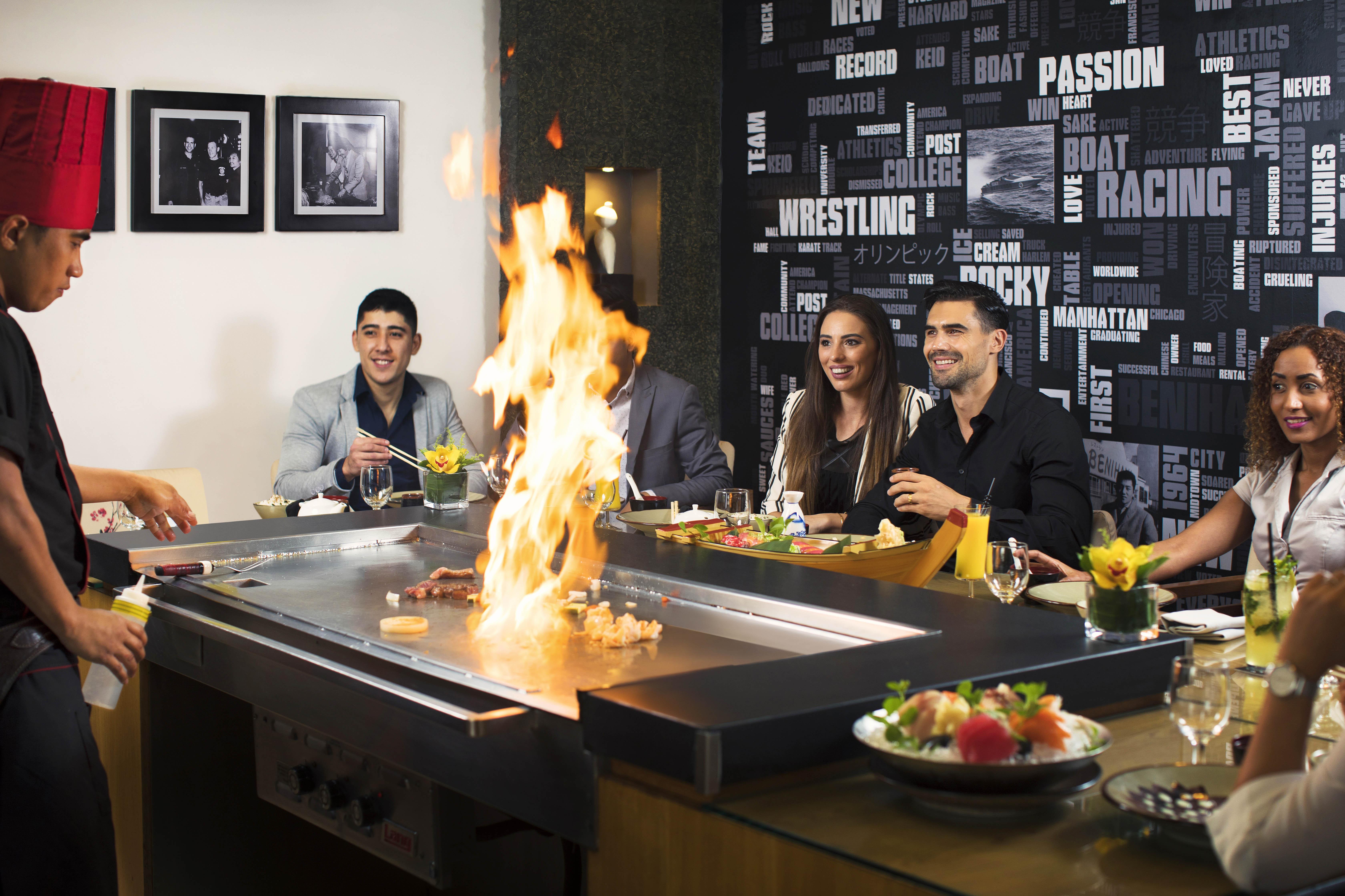 Amwaj Rotana, Jumeirah Beach - Dubai Esterno foto The photo shows a lively dining scene at a restaurant where hibachi cooking is taking place. In the foreground, a chef is preparing food on a grill, creating flames, which adds excitement to the experience. There are five diners seated around the gri