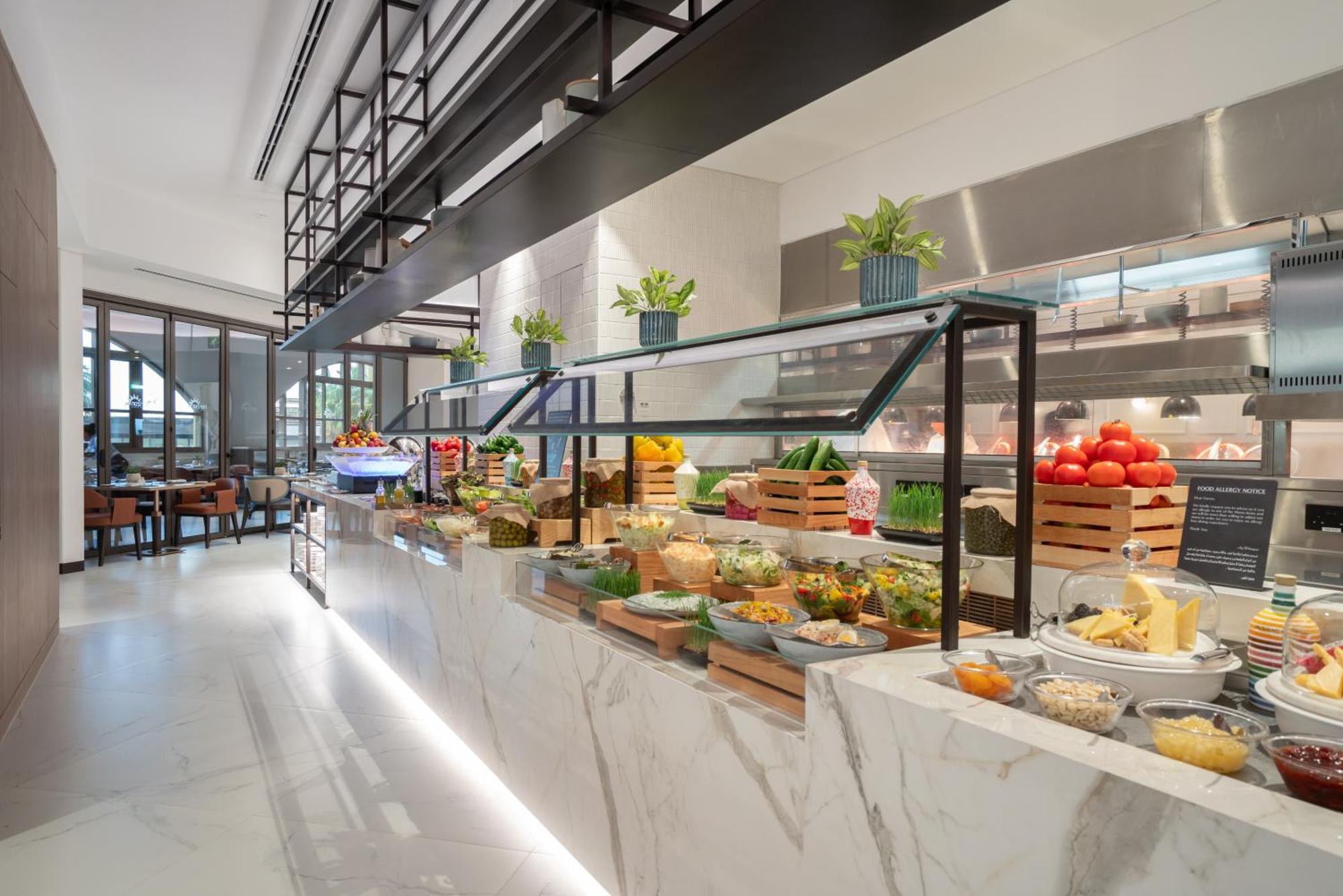 Amwaj Rotana, Jumeirah Beach - Dubai Esterno foto The photo depicts a modern, stylish buffet area in a restaurant. It features a long marble countertop with an array of fresh ingredients and food items displayed attractively. There are different types of vegetables, fruits, cheeses, and other dishes