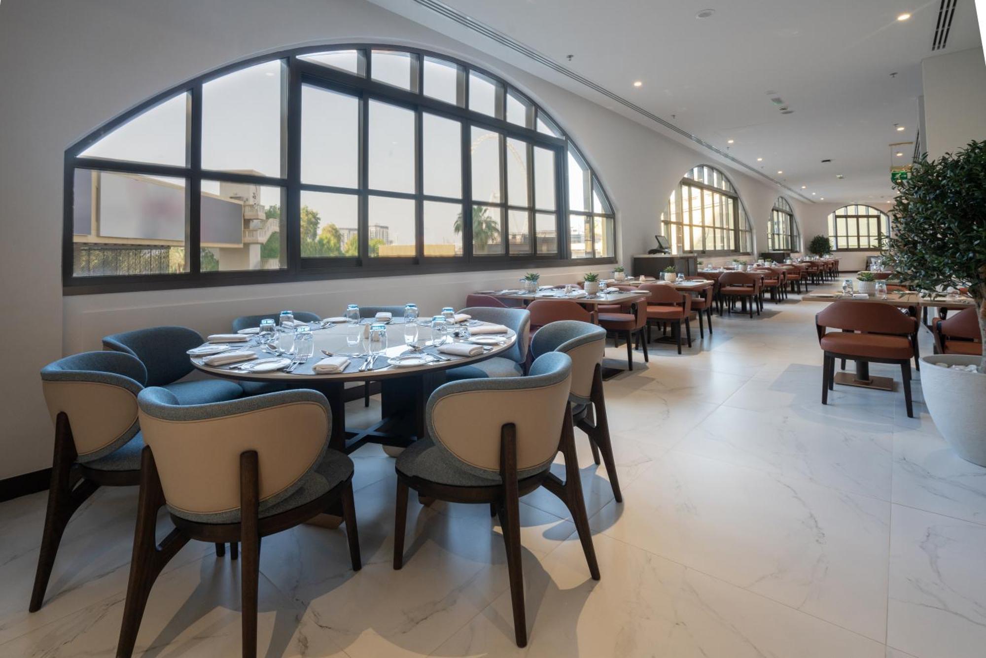 Amwaj Rotana, Jumeirah Beach - Dubai Esterno foto The photo shows the interior of a modern restaurant. It features a spacious layout with several tables arranged around the room. The design includes elegant, upholstered chairs with a mix of colors and round tables set with plates and cutlery. Large 