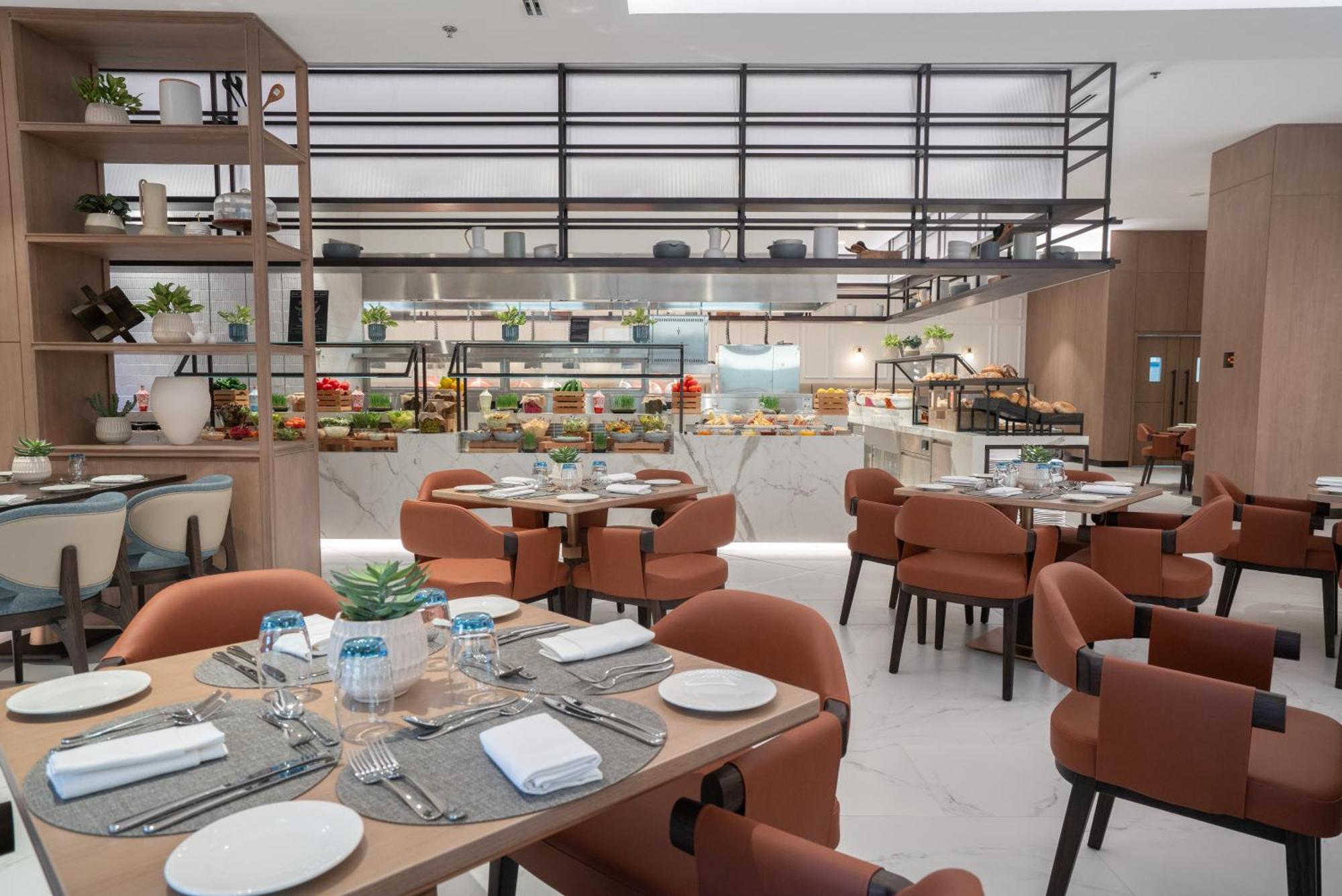 Amwaj Rotana, Jumeirah Beach - Dubai Esterno foto The photo shows a modern restaurant interior with a clean and stylish design. In the foreground, there are several tables set for dining, each with plates, cutlery, and glasses. The chairs are a warm rust color, creating a cozy atmosphere. 

In the b