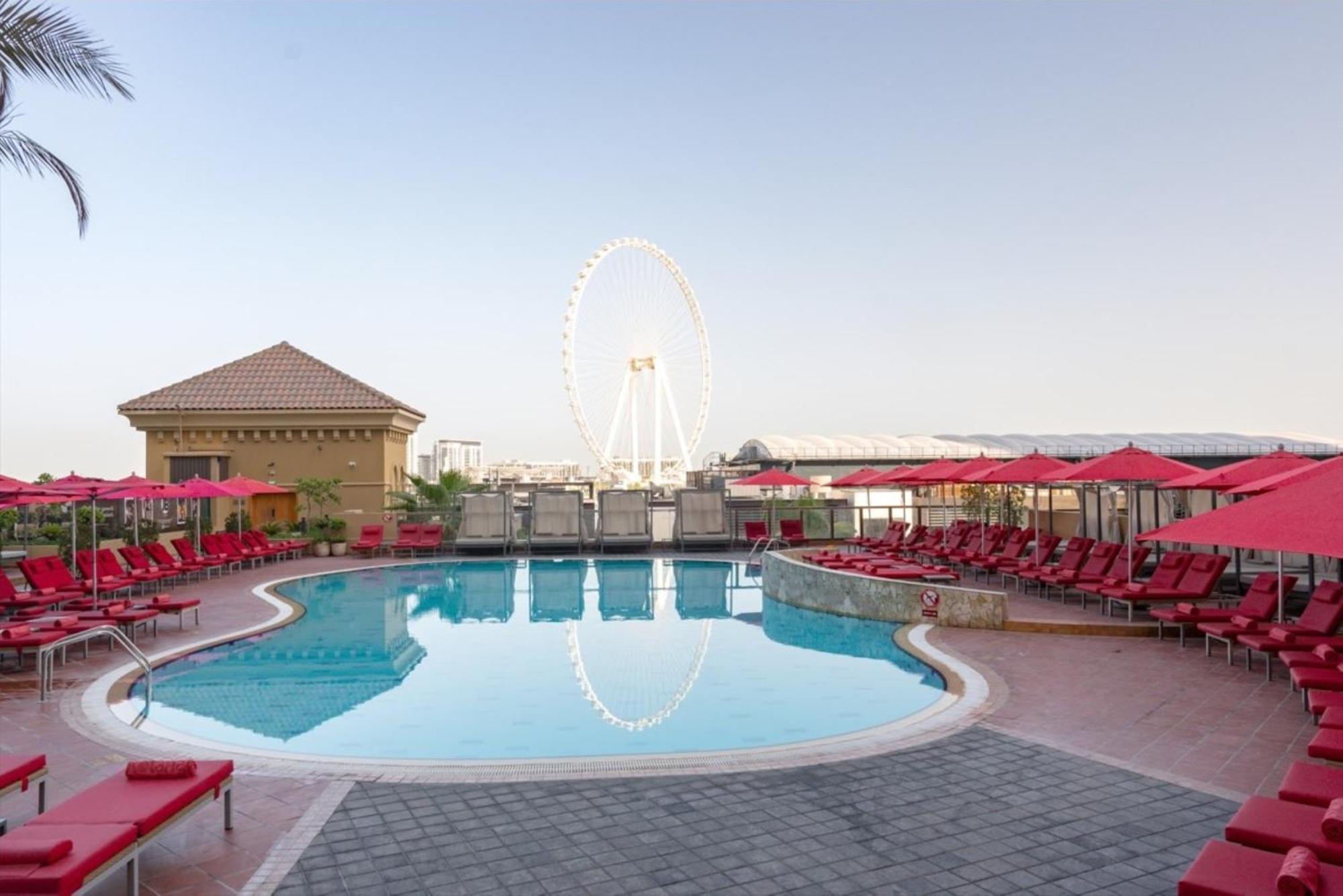 Amwaj Rotana, Jumeirah Beach - Dubai Esterno foto The photo depicts a swimming pool area with bright red lounge chairs and umbrellas surrounding the pool. In the background, there is a large Ferris wheel, indicating a nearby amusement or attraction. The setting appears to be outdoors, likely in a re
