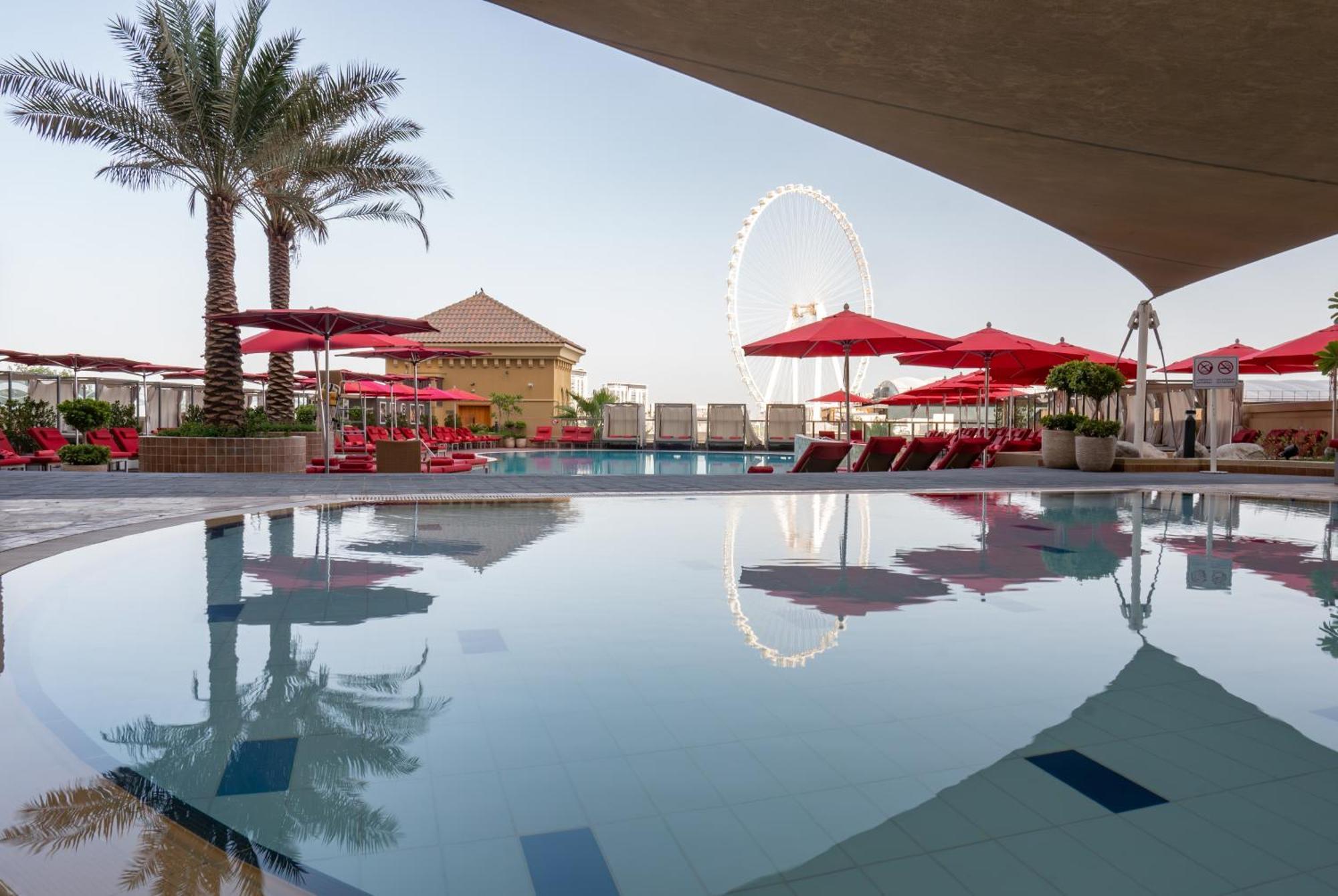 Amwaj Rotana, Jumeirah Beach - Dubai Esterno foto The photo depicts a luxurious outdoor pool area surrounded by palm trees and lounge chairs. There are several red umbrellas set up near the poolside, creating a vibrant and inviting atmosphere. In the background, there's a large wheel—likely a Ferris