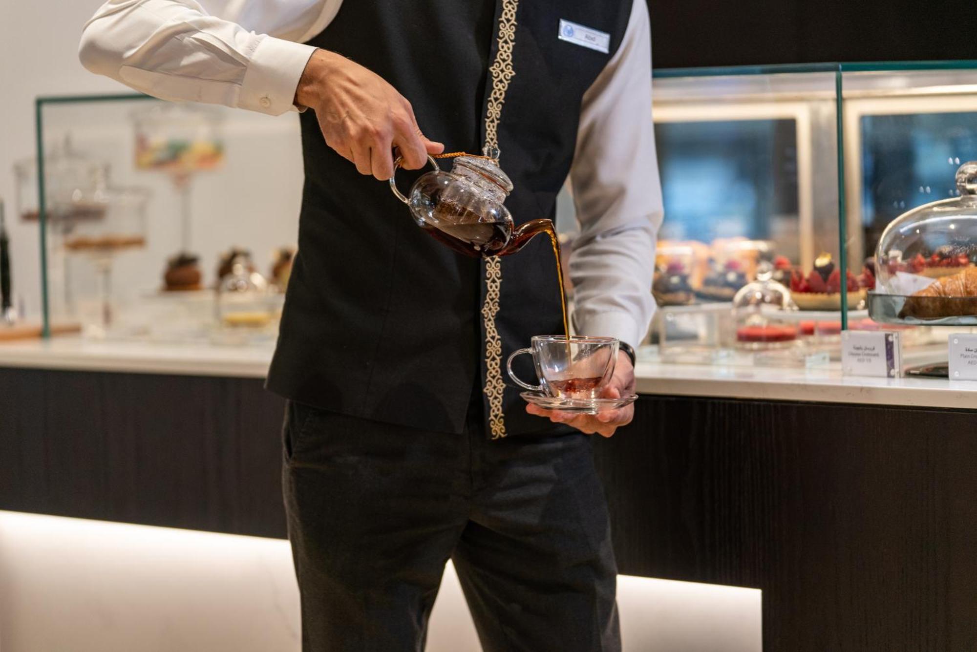 Amwaj Rotana, Jumeirah Beach - Dubai Esterno foto The photo shows a person in a black vest and white shirt pouring a beverage from a glass teapot into a clear glass cup. The setting appears to be a café or restaurant, with a display case in the background containing various pastries or desserts. The