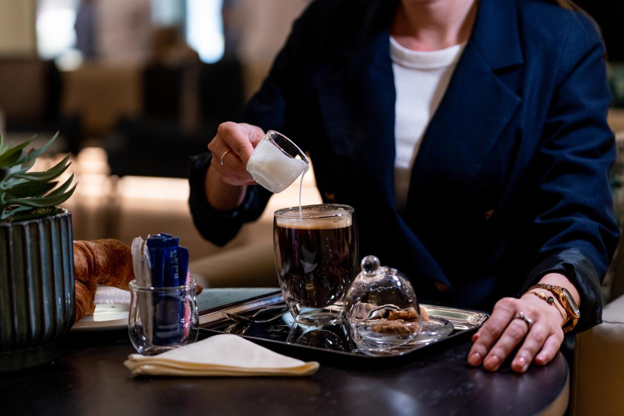 Amwaj Rotana, Jumeirah Beach - Dubai Esterno foto The photo shows a person pouring cream from a small pitcher into a cup of coffee. The coffee is on a tray, which also holds a glass dome covering some pastries or cookies. There are utensils in a holder, a folded napkin, and a croissant visible on th