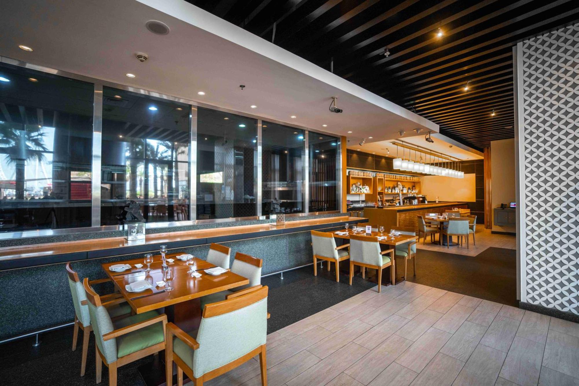 Amwaj Rotana, Jumeirah Beach - Dubai Esterno foto The photo shows the interior of a modern restaurant. In the foreground, there are several wooden tables set with utensils, plates, and glassware. The chairs are upholstered in a light fabric. The background features a counter area with a coffee machi