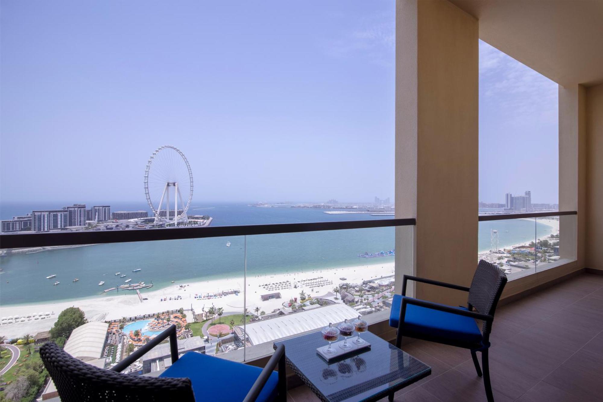 Amwaj Rotana, Jumeirah Beach - Dubai Esterno foto The photo depicts a balcony scene overlooking a beach and sea. There's a modern, stylish balcony with two blue chairs and a small glass table. In the background, you can see a wide expanse of sandy beach, water, and a large Ferris wheel. The sky is c